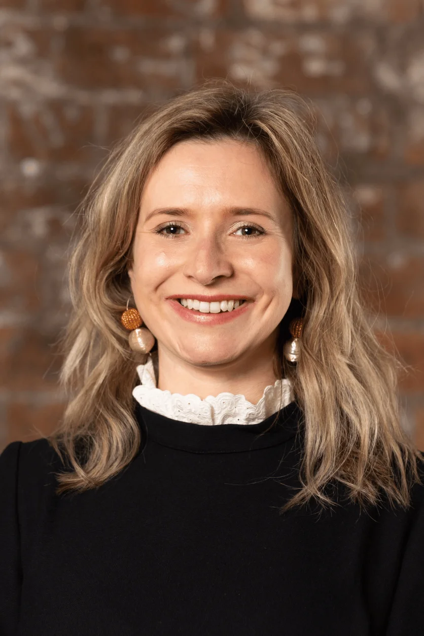 Photo of a white woman with brown shoulder length hair. She wears a black top and white collar.