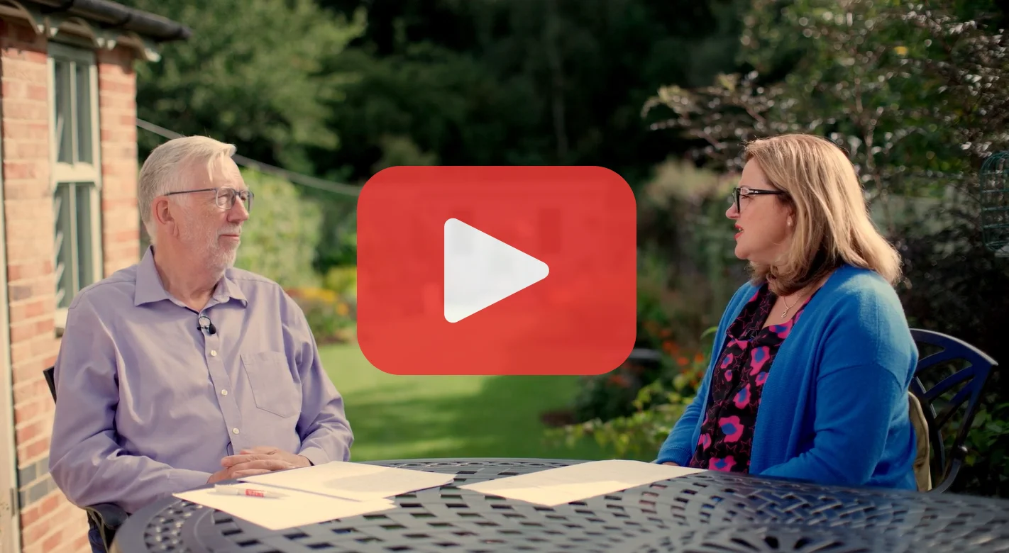 Photo shows a white man sitting at a table in a garden with a white woman. Both are looking at eachother and in conversation.. The photo has a play button overlaid.