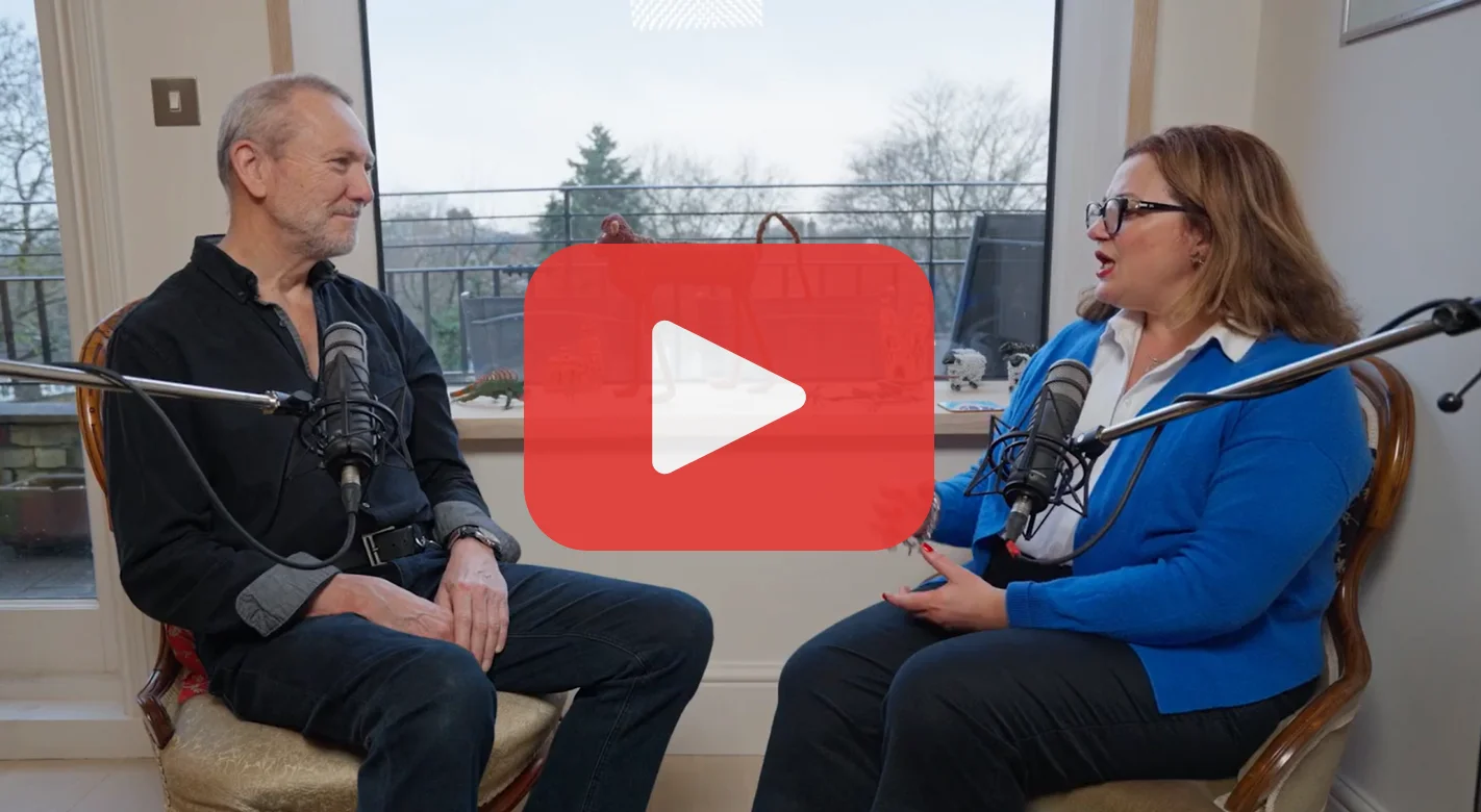 Photo shows a white man and white woman sitting next to each other with microphones propped next to them. There is a play button overlaid.